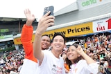 A man smiles with two others while posing for a selfie on stage in front of a large crowd.