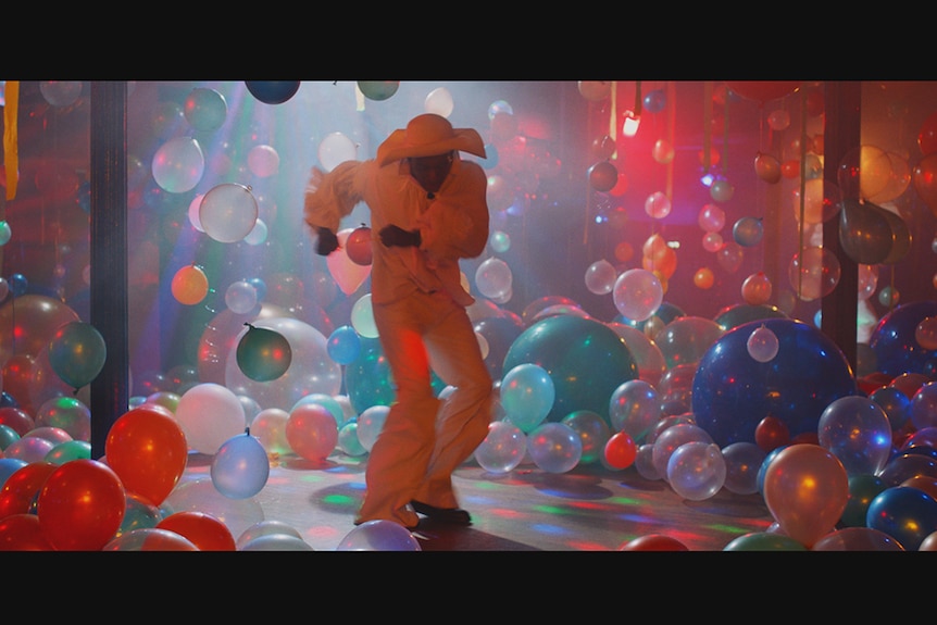 Colour still of a person dressed in all white dancing in a room full of balloons and colour lights.