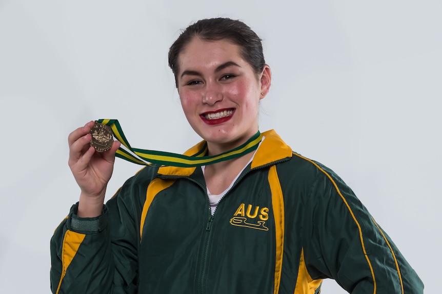 Ada Lacey, a young woman, wears an Australian tracksuit and holds up a gold medal 