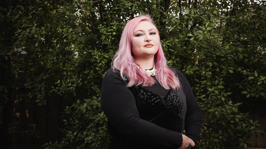 A woman with pink hair, wearing a black dress, looks stoicly towards the camera with dark green foilage behind her. 