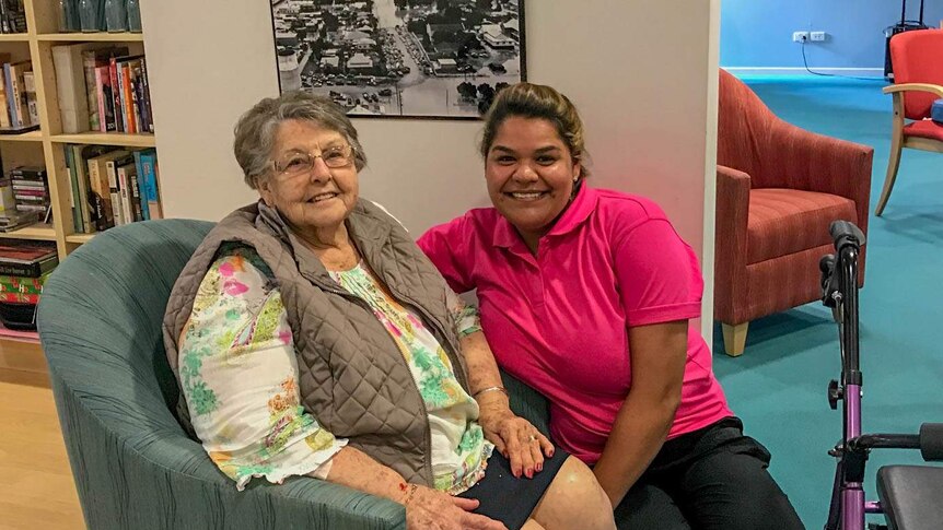 An elderly woman sitting in a chair at a nursing home beside an indigenous carer