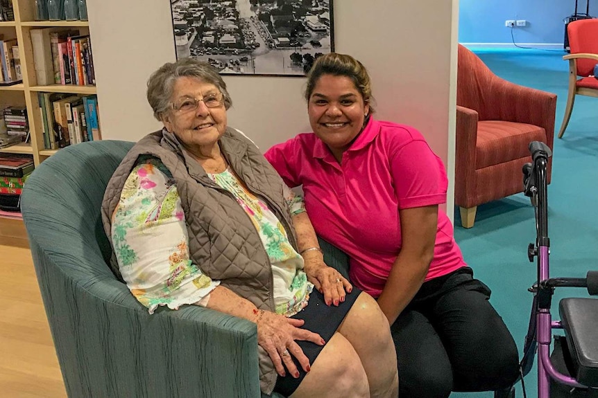 An elderly woman sitting in a chair at a nursing home beside an indigenous carer
