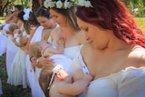 Women dressed in white breastfeed their babies in a field with trees