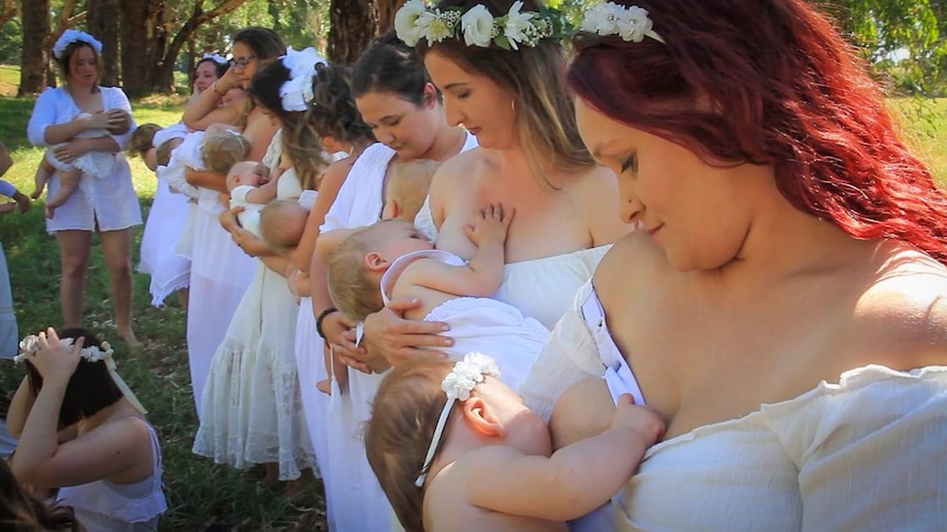 Women dressed in white breastfeed their babies in a field with trees