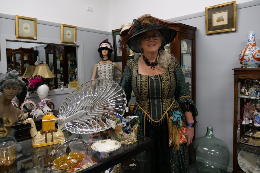 A woman in a green and gold vintage stands with her antique wares. 