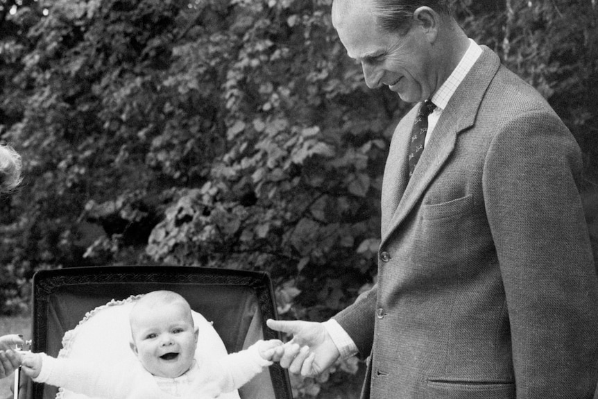 A black and white photo of Prince Philip in a suit holding the hand of baby Prince Andrew in a pram.