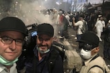 Lipson, cameraman and producer standing in front of crowd with tear gas in background.