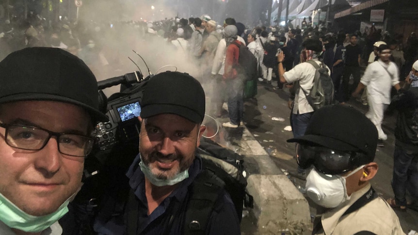 Lipson, cameraman and producer standing in front of crowd with tear gas in background.