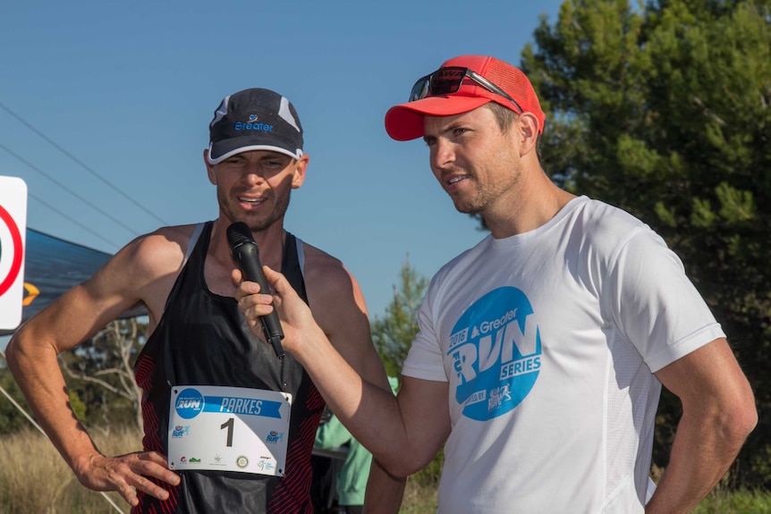 A man in a hat and running singlet with another man holding a microphone to the first man's mouth