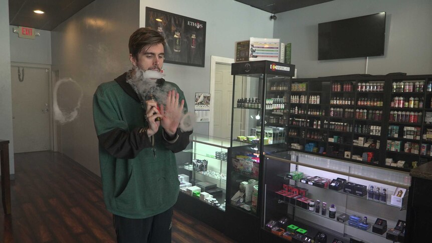 Justin Brooks stands in a vape shop as he blows smoke rings from a vape. He wears a green jumper and dark track pants.