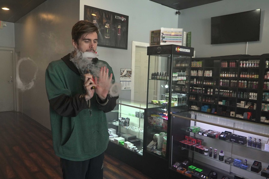 Justin Brooks stands in a vape shop as he blows smoke rings from a vape. He wears a green jumper and dark track pants.