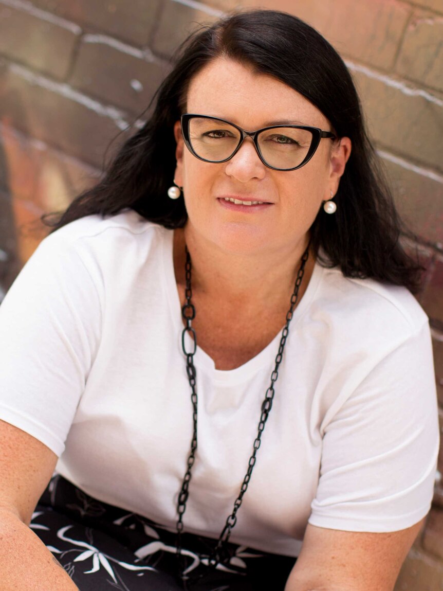 A woman sits in front of a brick wall looking into the camera. She wears glasses and pearl earrings.