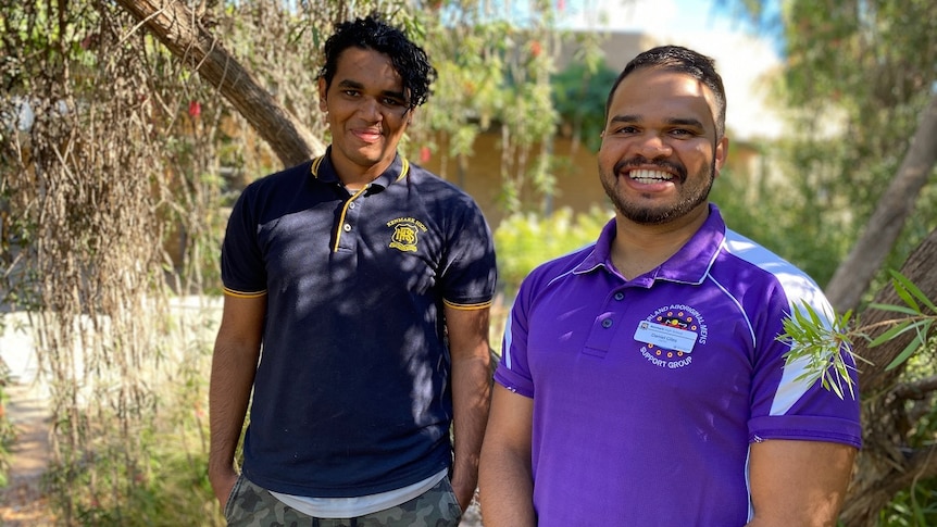 Two men are standing smiling at the camera. There is a tree and blurry building behind them.