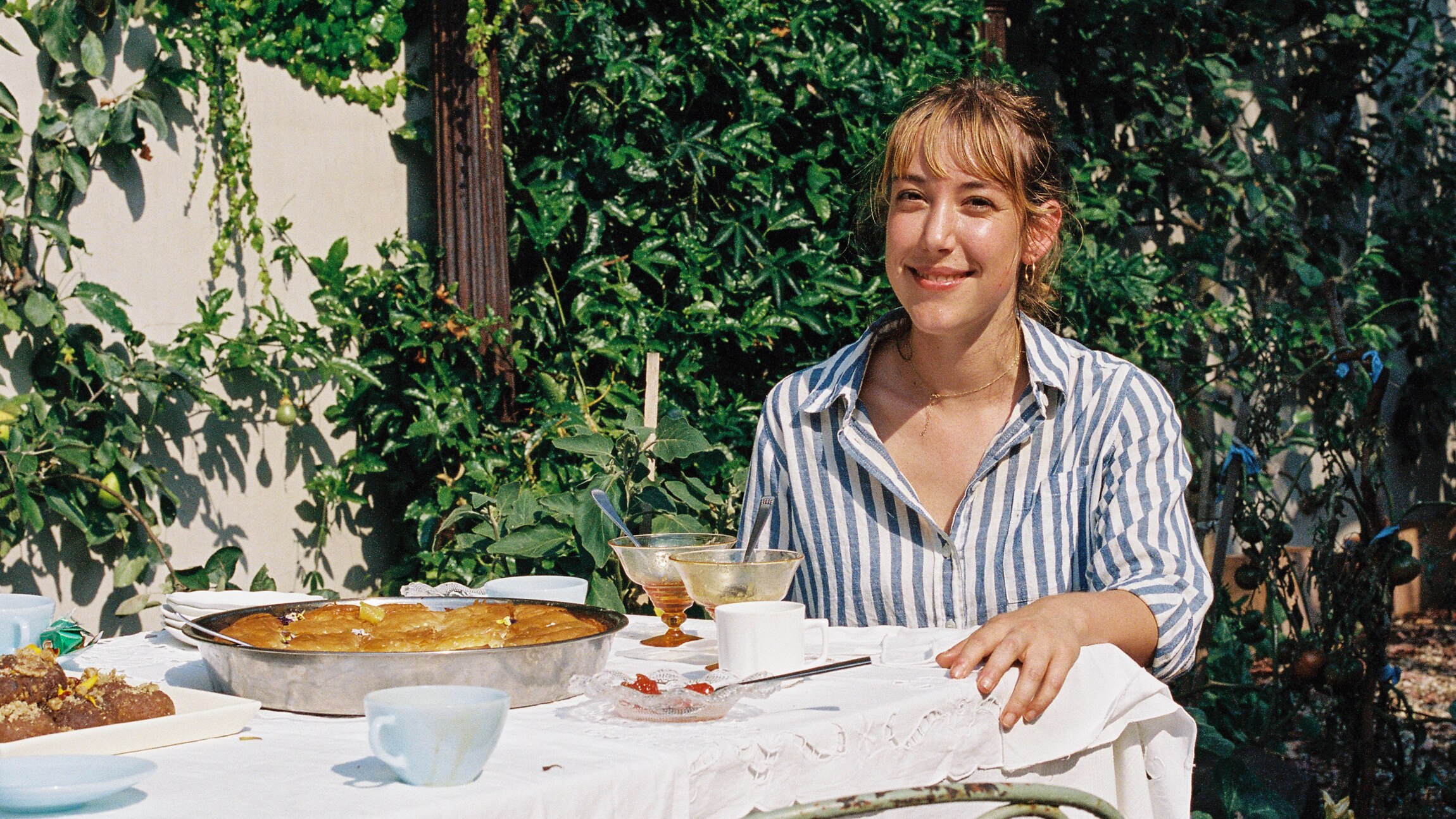 Ella sits at an outdoor table covered with greek sweets, she is wearing a blue and white shirt and there are vines behind her