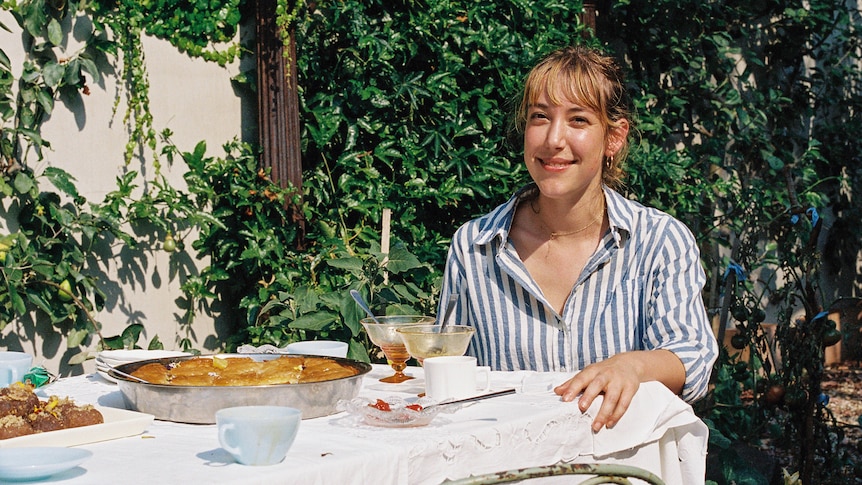 Ella sits at an outdoor table covered with greek sweets, she is wearing a blue and white shirt and there are vines behind her