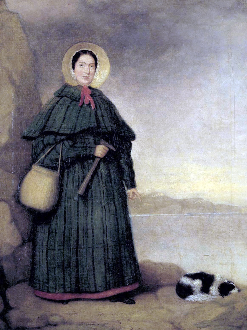 A portrait of Mary Anning wearing a cloak, bonnet and carrying a pick and basket while standing on the beach.