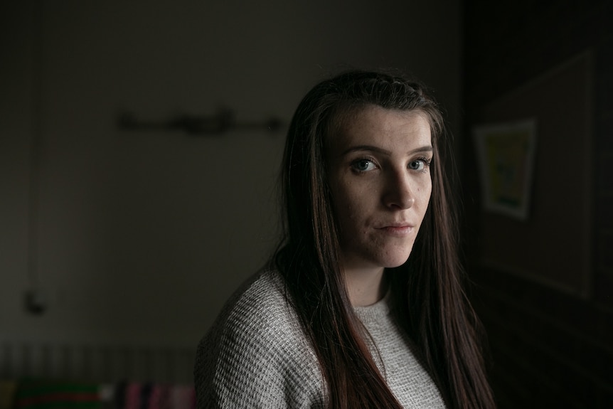 A woman standing in a dark room, with light hitting the side of her face, looks at the camera with a neutral expression.