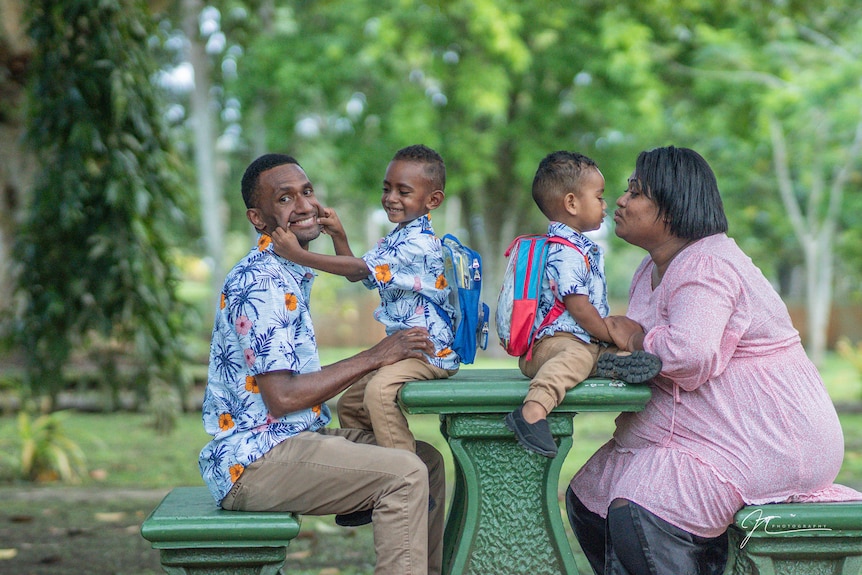 Two small boys sit on green table in parkland. One boy kisses his Mum. The other boy pulls his Dad's face. 