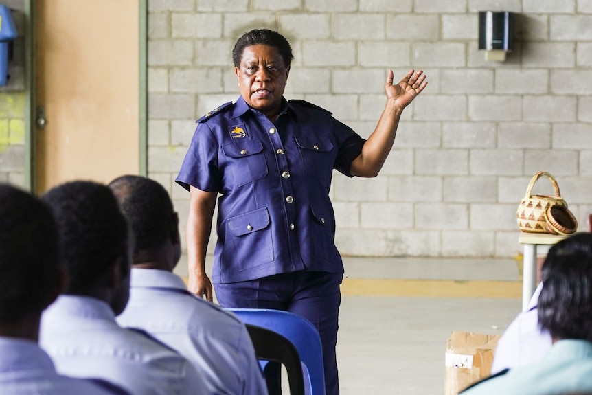 A woman wearing a navy uniform stands in front of a group of people pointing to the side.