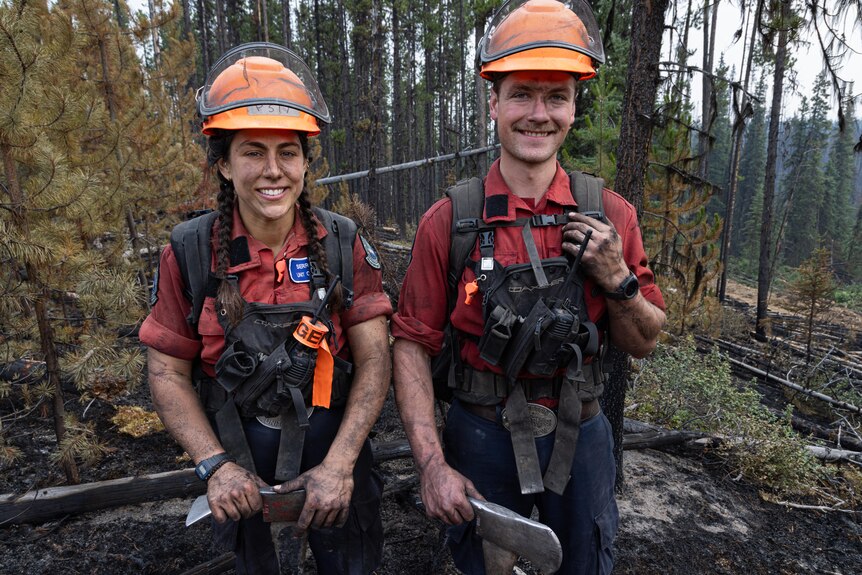 Two young firefighters, one female and one male, stand in a forest smiling. Their skin is ashened and they wear helmets.