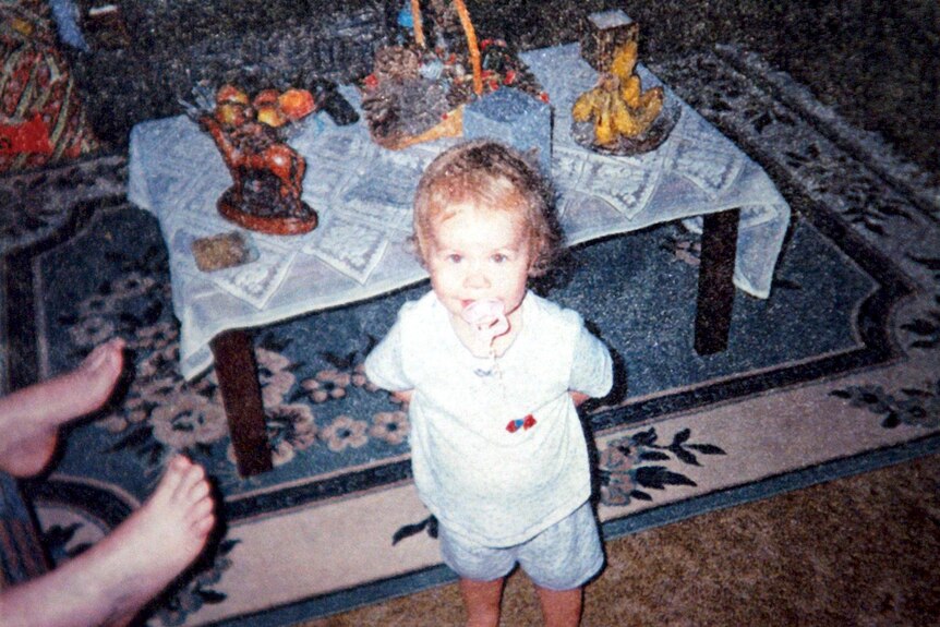 A young girl with a dummy in her mouth stands in a living room in front of a decorated coffee table