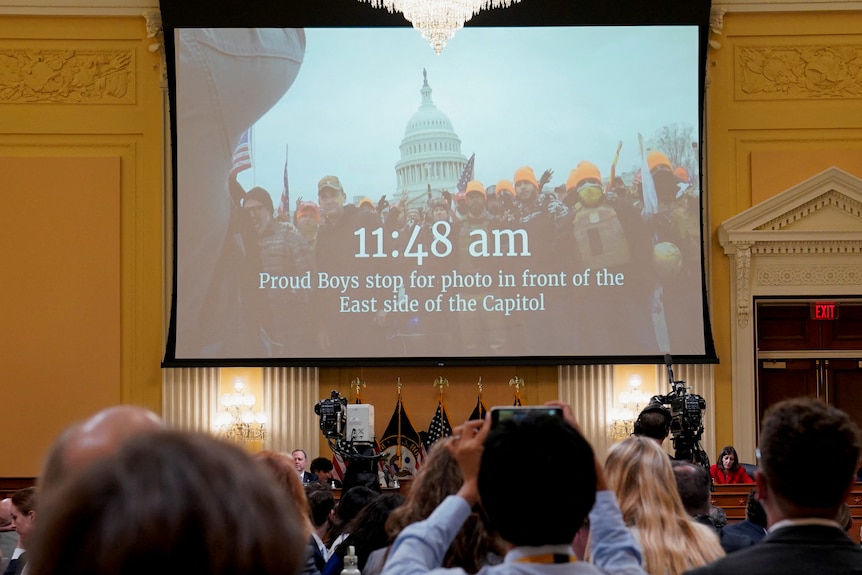 a group of people watch images of the capitol riots on a large drop-down projector screen during a us senate committee hearing
