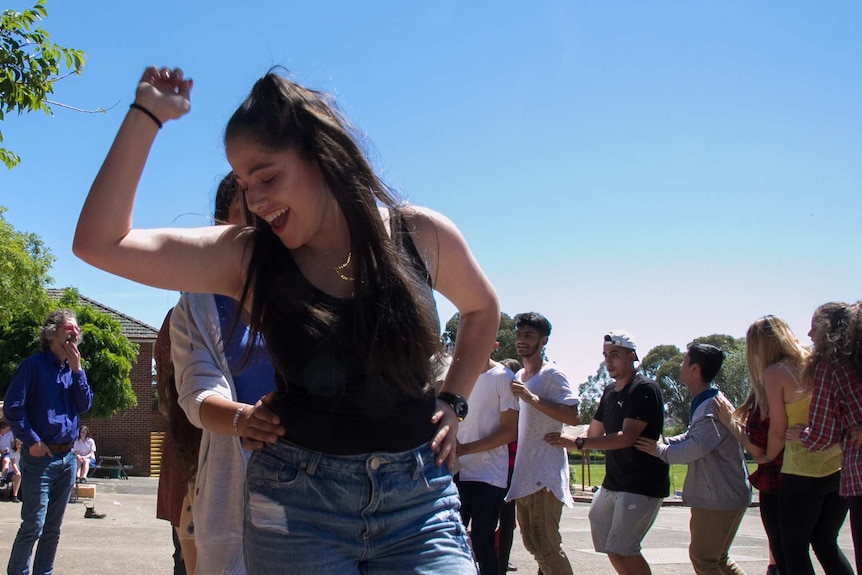 A teenaged girl leading a conga line of young people dancing in a school playground.