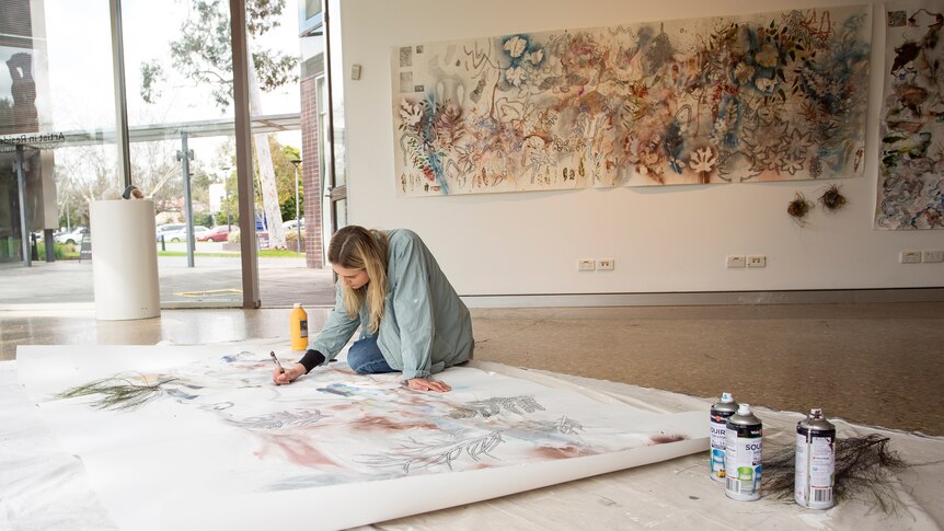 An artist sits on a gallery floor creating art. On the wall behind her is a large canvas painting.