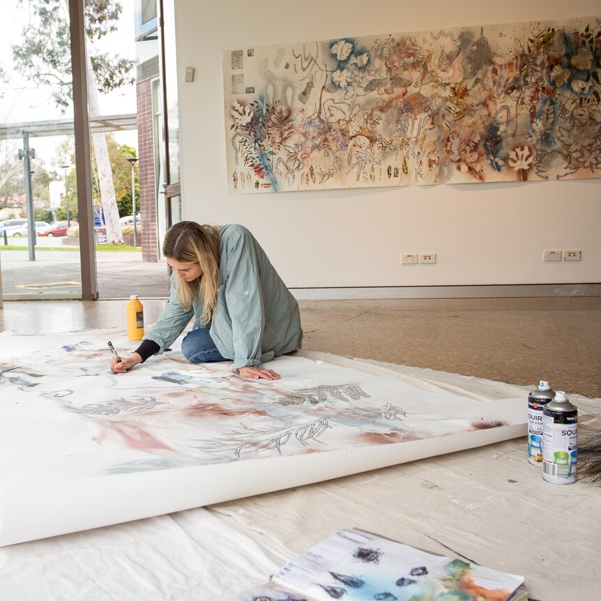 An artist sits on a gallery floor creating art. On the wall behind her is a large canvas painting.