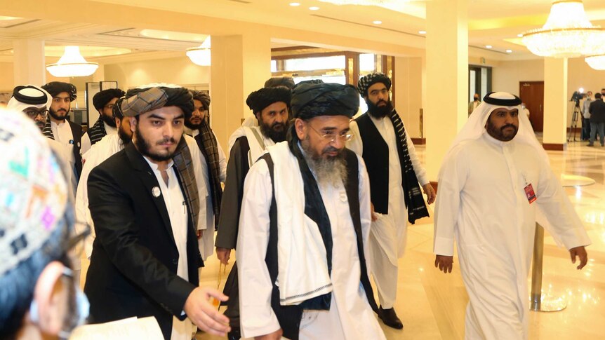A group of men wearing traditional Afghan clothing walk through a brightly lit lobby.