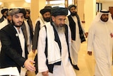 A group of men wearing traditional Afghan clothing walk through a brightly lit lobby.