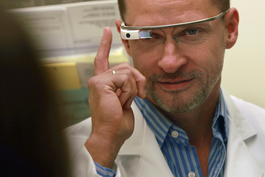 A middle-aged white man in a lab coat touches the side of his Google Glass glasses