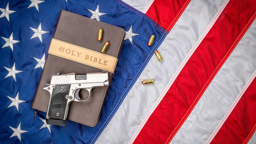 Handgun placed on a copy of the Holy Bible, with bullets scattered around it, both gun and Bible sitting on the US flag.