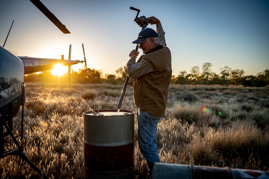 A man is using a long steel implement to pour fuel into a steel barrel, next to a helicopter at the crack of dawn.
