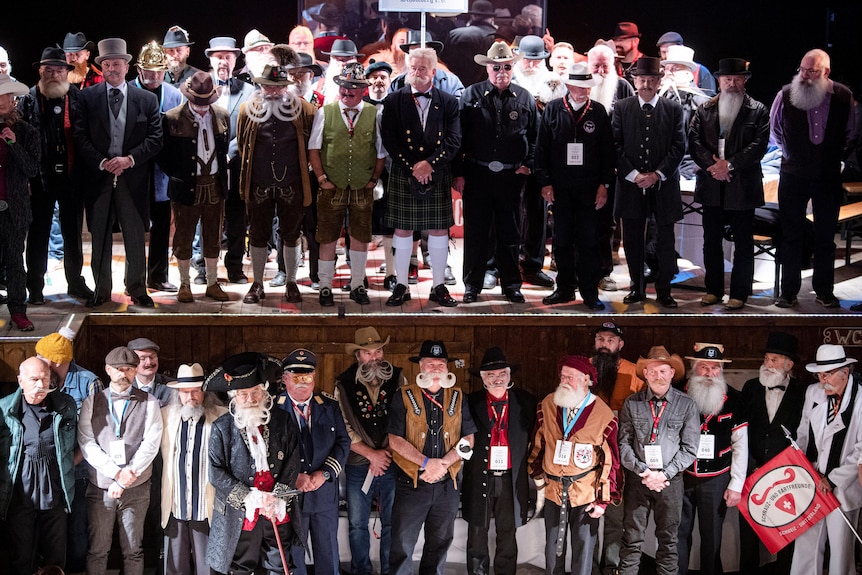 A stage of men stand in fancy dress showcasing their perfectly manicured white beards