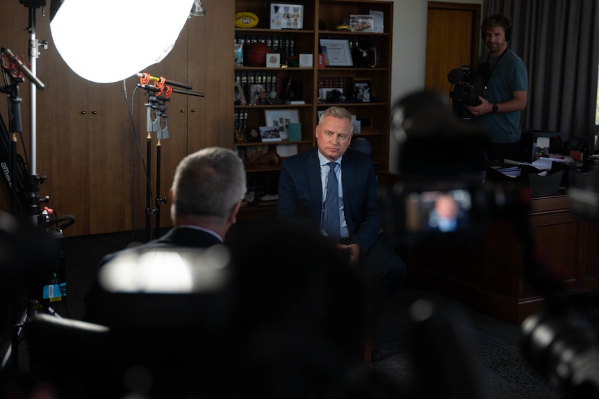 A male politician sits in a suit during an interview with a journalist surrounded by three cameras and a light.