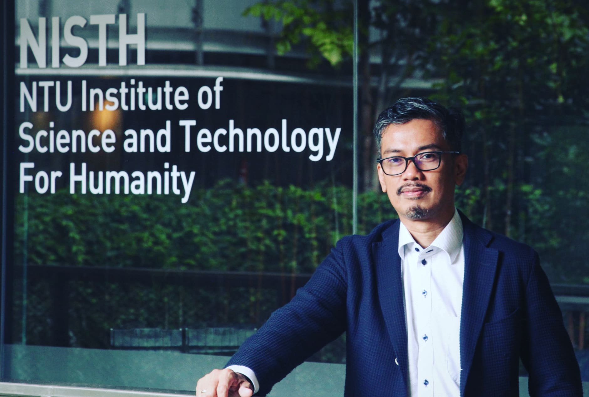 Man wearing suit and glasses facing the camera in front of the NTU Institute of Science and Technology sign.