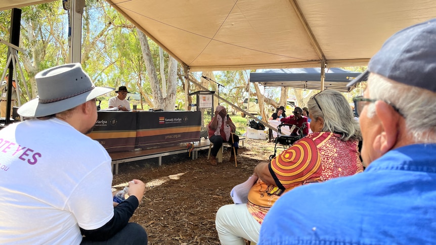 People under a marquee in the bush watch a man speaking at a podium.