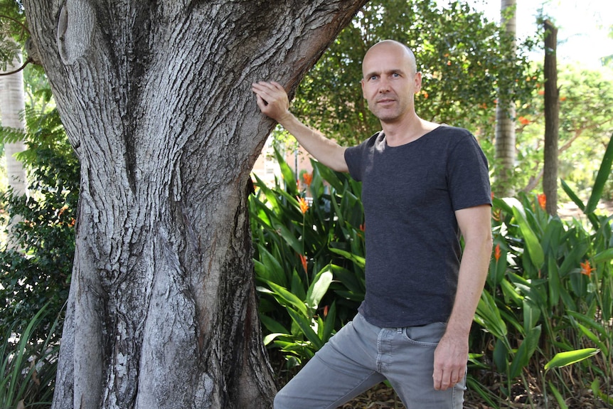 A young man in black jeans stands leaning against a tree in a tropical setting.