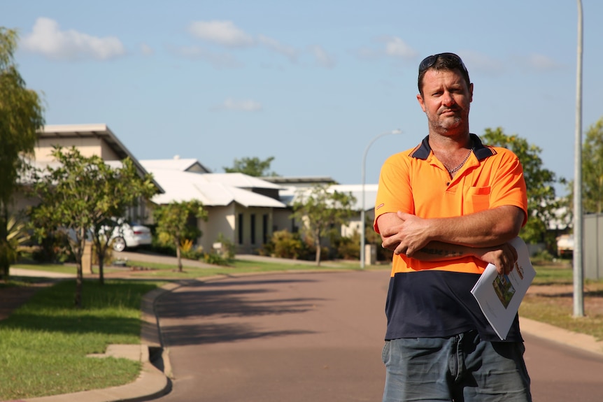 A man in an orange high-visibility shirt stands in a street with arms folded, holding a report.