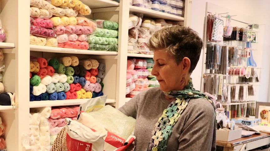 An older woman wearing a knitted scarf holds a basket of wool in a yarn store.