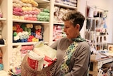 An older woman wearing a knitted scarf holds a basket of wool in a yarn store.