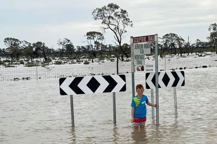 Boy stands in floodwaters at road signs at cut Jericho Road near Aramac.