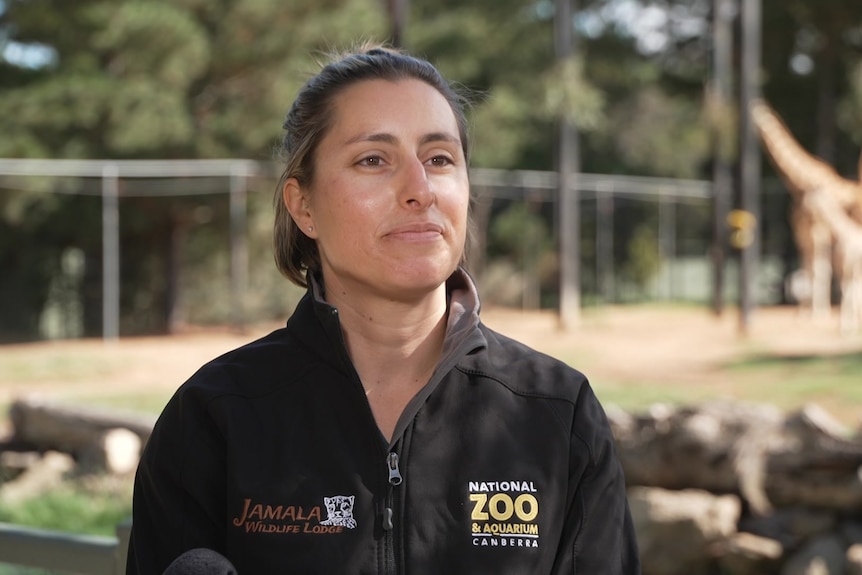A woman in a zoo branded polo smiles in front of a giraffe enclosure.