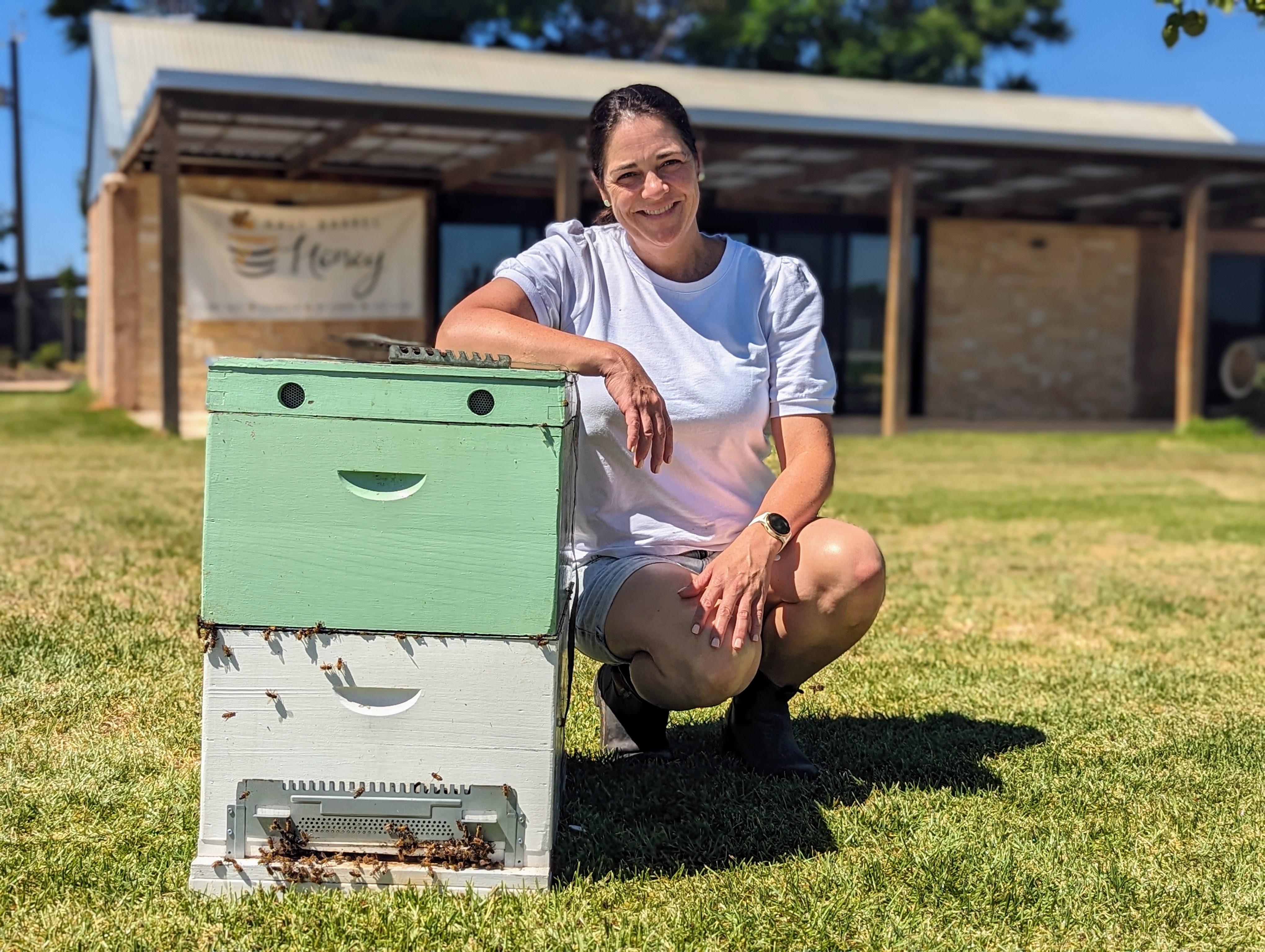 A fair-skinned brunette woman, Kerry, kneels beside green and white hives in full sun.