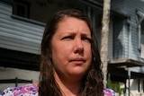 Woman in purple dress stands outside flood-impacted home and caravan 