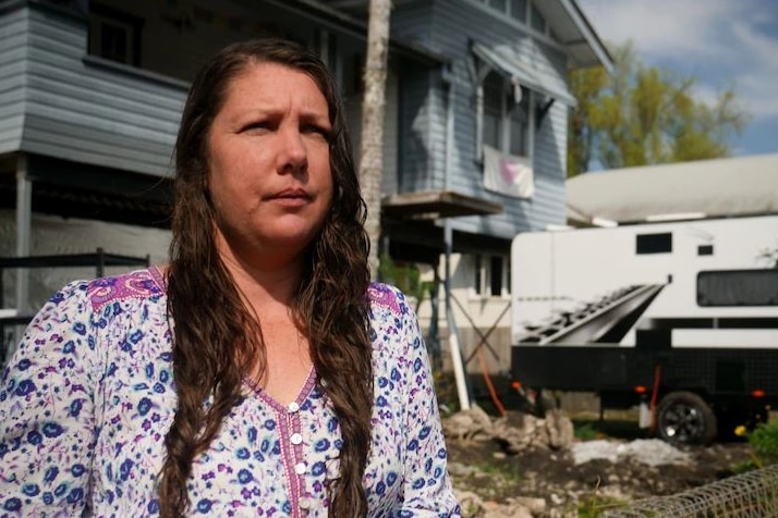 Woman in purple dress stands outside flood-impacted home and caravan 