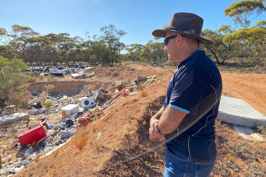 A man is standing on brown soil looking over a hole full of rubbish. There are lots of trees in the background.