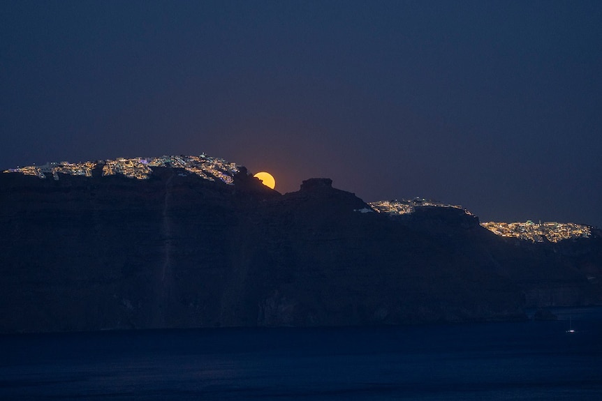 the moon peeks out from the skyline of a village in santorini 