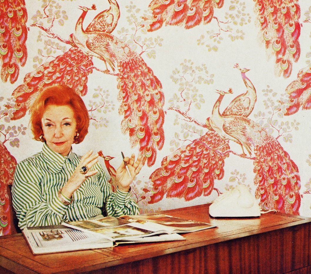 A woman with short red hair sits behind a desk, with books on it, in front of peacock-prinyed wallpaper. 